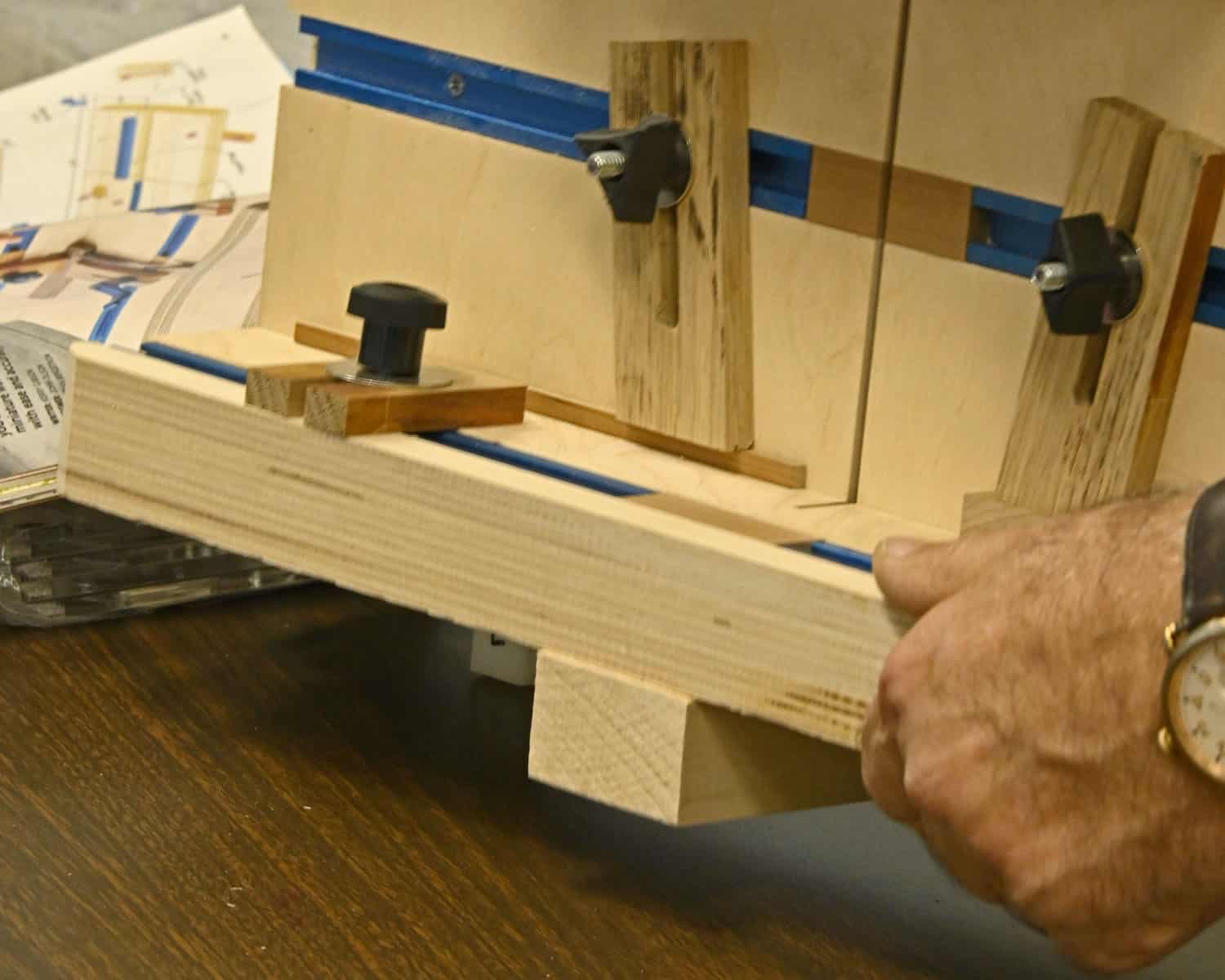 A member's hand holding a shop-built jig alongside its measured drawings on the table.