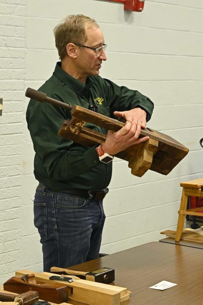 A member holds an antique wooden plow plane with wooden screw arms.